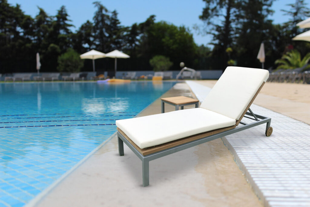 Celeste Resort teak sun lounger for hotels beside a pool with matching side table and rear wheels visible
